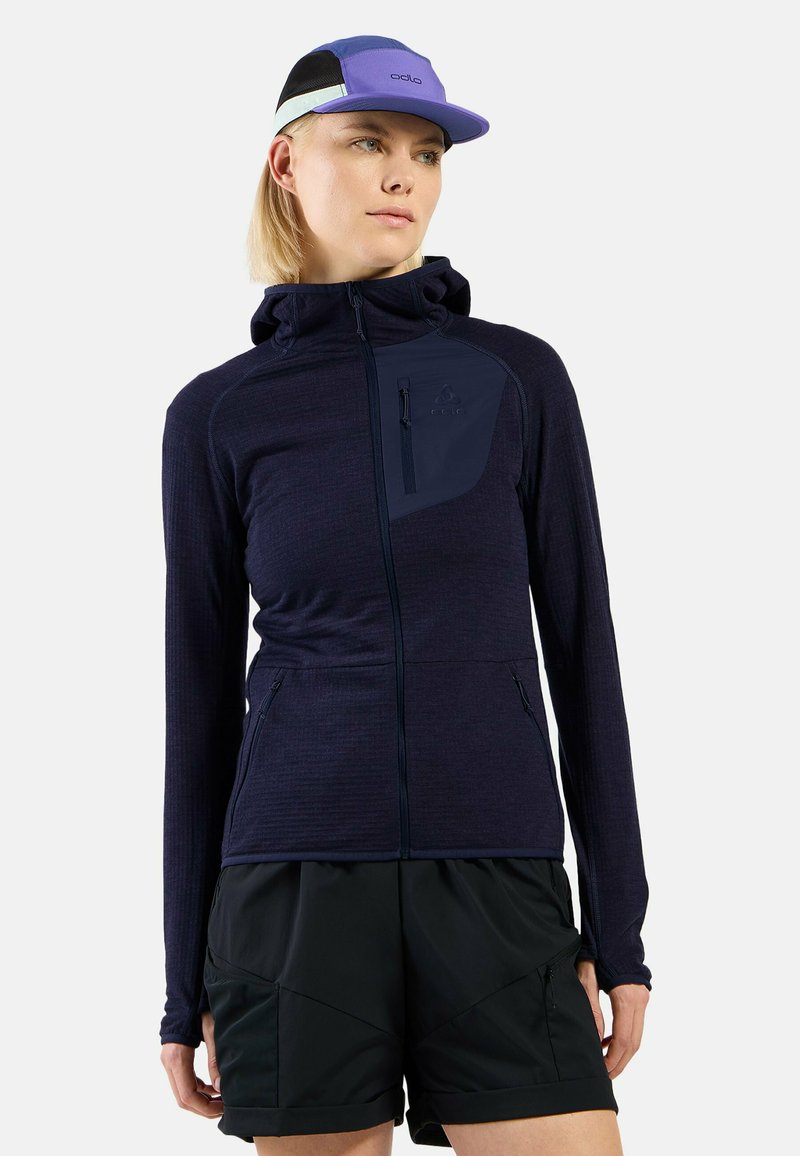 Navy Zip-Up-Jacke mit Kapuze und strukturiertem Stoff, mit einer kontrastierenden dunklen Tasche und hohem Kragen, kombiniert mit schwarzen Shorts.