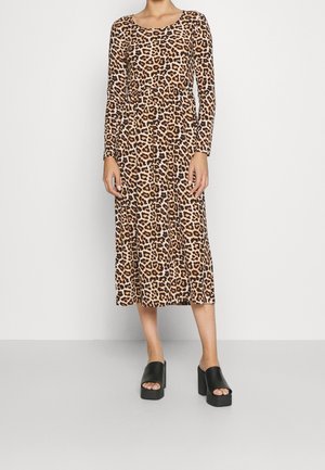 Mujer con vestido de manga larga hasta el tobillo con estampado de leopardo y zapatos negros de plataforma y punta abierta, de pie frente a un fondo liso.