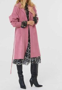 Femme portant un long manteau rose sur une robe à fleurs avec un ourlet imprimé léopard, associée à des bottes noires à talons jusqu’aux genoux, debout sur un fond blanc.