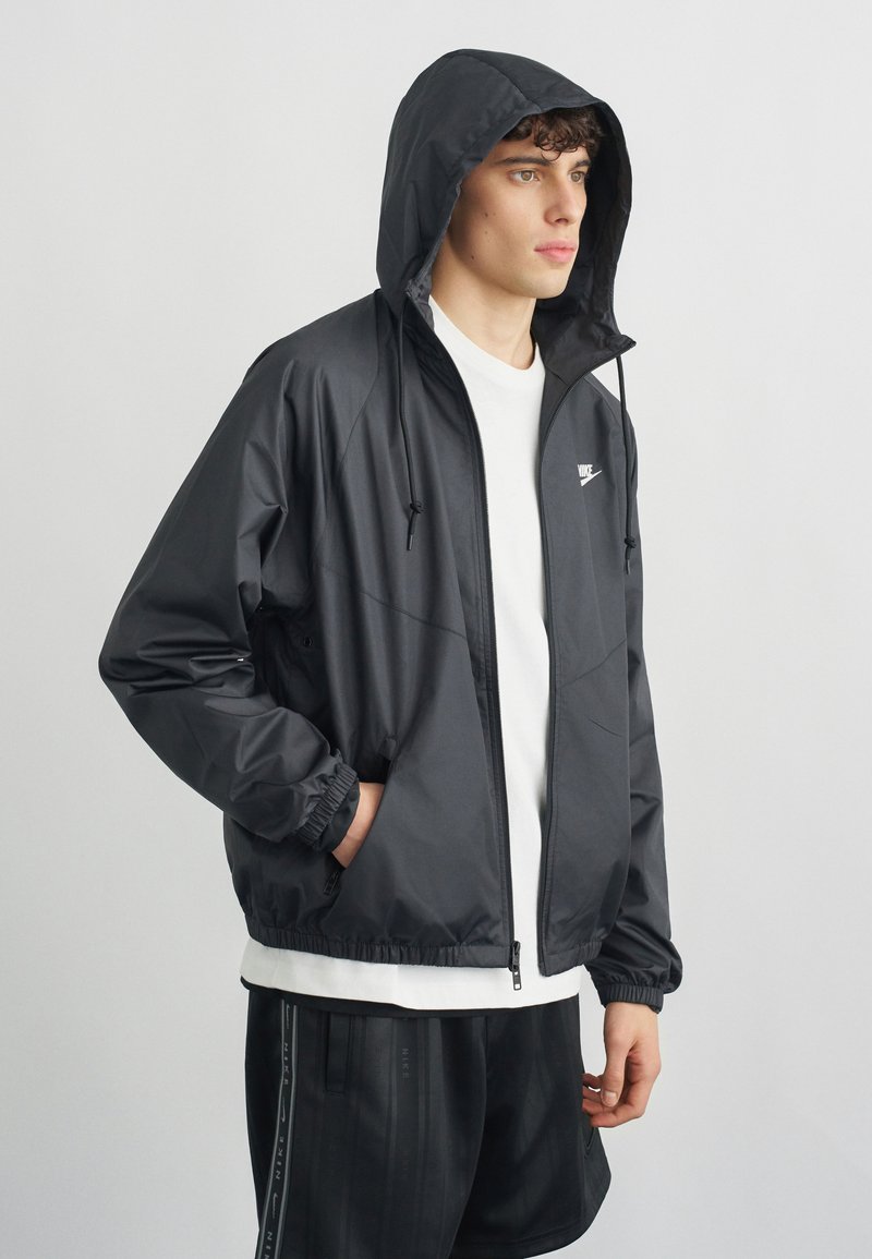 Giovane uomo con giacca a vento con cappuccio Nike nera, t-shirt bianca e pantaloncini neri, in piedi di profilo verso destra con le mani nelle tasche contro uno sfondo semplice.