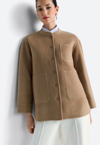 Beige, gestructureerd fleecejack met een ronde hals, knopen aan de voorkant en twee grote zakken. Gedragen over een blauw-wit gestreept shirt.