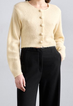 Femme portant un cardigan duveteux crème boutonné et un pantalon noir taille haute, une main dans la poche, sur un fond clair uni.