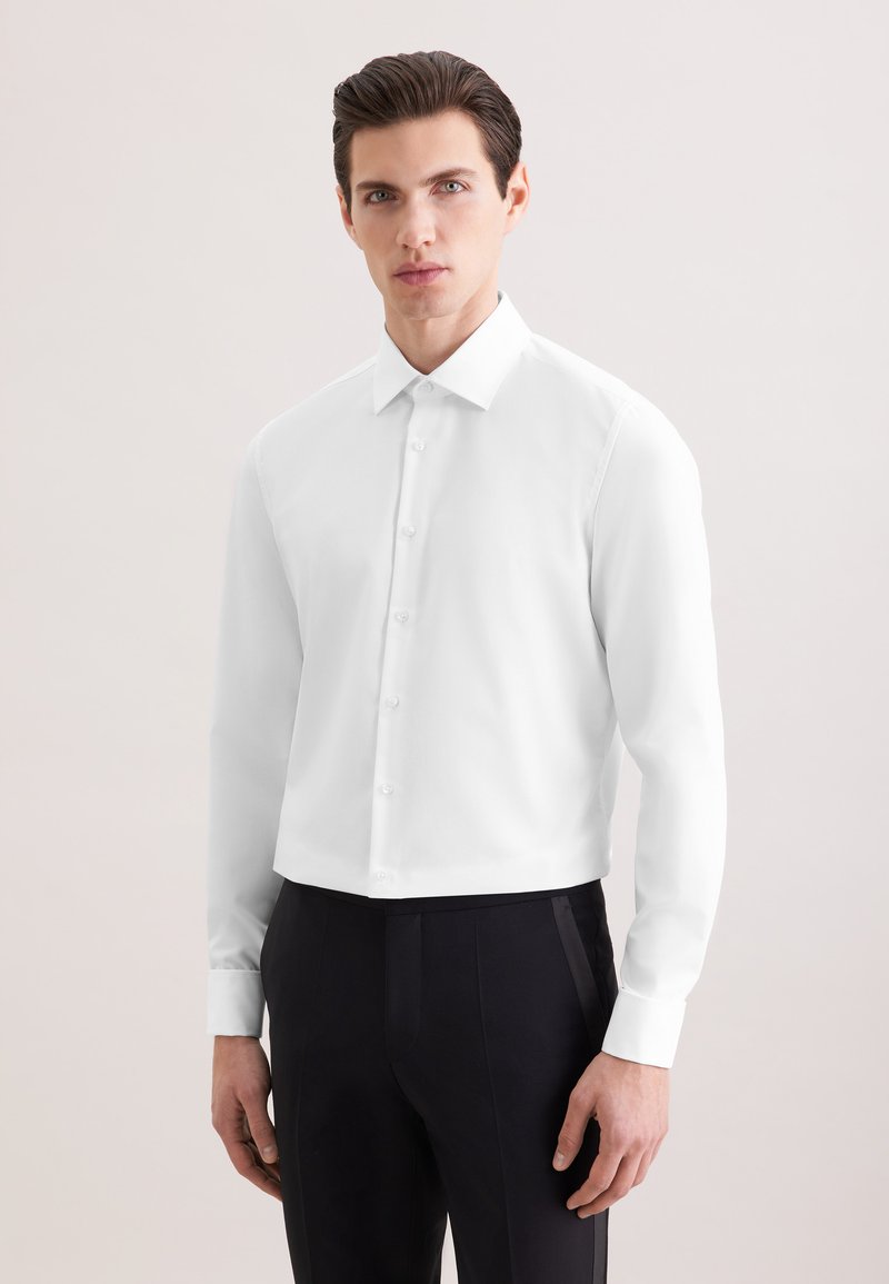 Jeune homme aux cheveux foncés portant une chemise blanche à manches longues et un pantalon noir, debout devant un fond clair uni.
