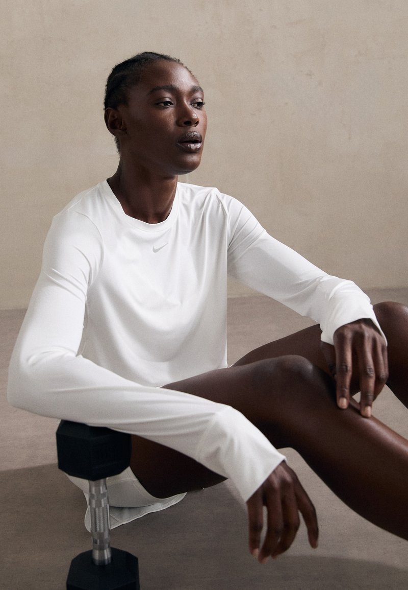 Long-sleeve white athletic shirt with a round neckline and Nike logo, paired with black dumbbell on textured surface.