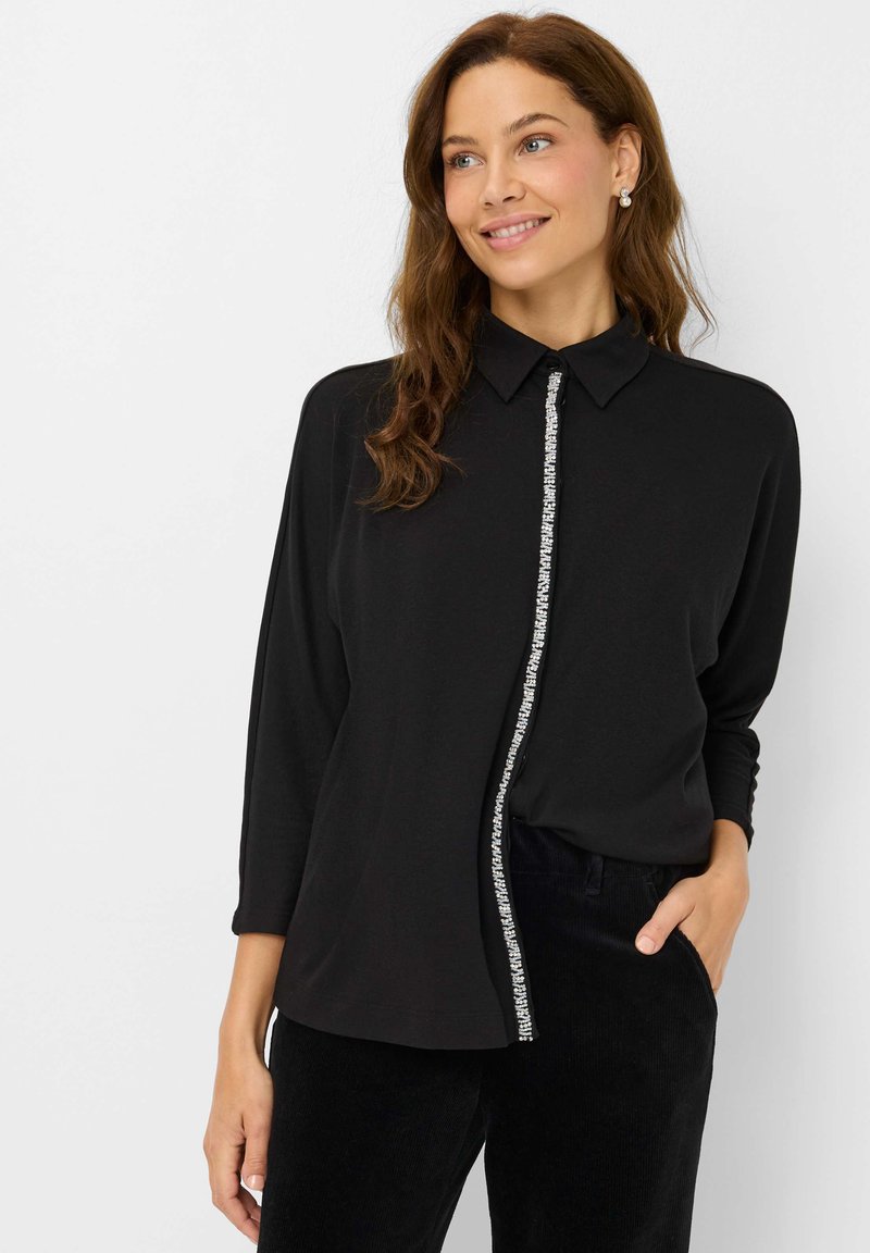 Frau in einem schwarzen Langarmshirt mit Kragen und einem dekorativen silbernen Reißverschluss, die lächelt und mit einer Hand in der Tasche steht.