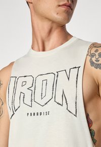 Sleeveless, light beige cotton tank top with "IRON" graphic in bold black letters. Features a round neckline and exposed shoulder design.