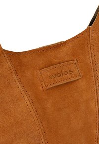 Brązowy zamszowy botek za kostkę, z logo na skórzanej naszywce i widocznymi detalami szwów, z okrągłym noskiem i gładką teksturą.