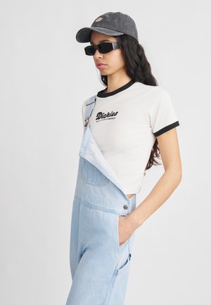 Jeune femme portant des lunettes de soleil noires, une casquette grise, un t-shirt blanc Dickies et une salopette en jean bleu clair avec une bretelle détachée.