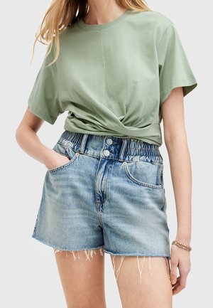 Mujer con camiseta de manga corta verde salvia metida en pantalones cortos de mezclilla azul claro de talle alto, con dobladillos deshilachados y cierre de doble botón.