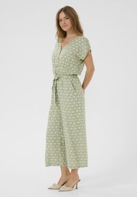 Jumpsuit vert à motifs avec des manches courtes, une taille nouée et des jambes larges. Le tissu semble léger avec une texture lisse.