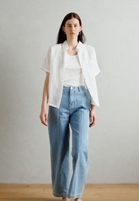 Camisa branca de manga curta, com botões, sobre uma t-shirt branca, combinada com jeans de cintura alta, azul claro. Design simples, corte descontraído.