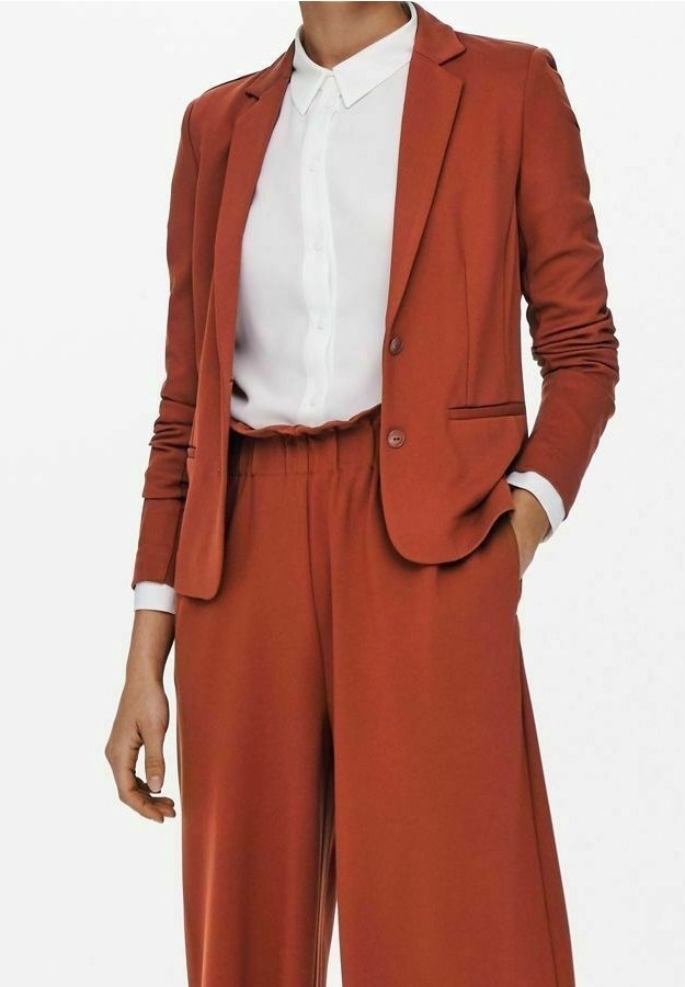 Femme portant un blazer couleur rouille et un pantalon taille haute à jambes larges assorti, avec une chemise blanche boutonnée rentrée dedans.
