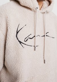 Beige, flauschiger Kapuzenpullover mit einem strukturierten, fleeceartigen Material. Verfügt über ein schwarzes besticktes Design und silberne Akzente an den Schnüren.