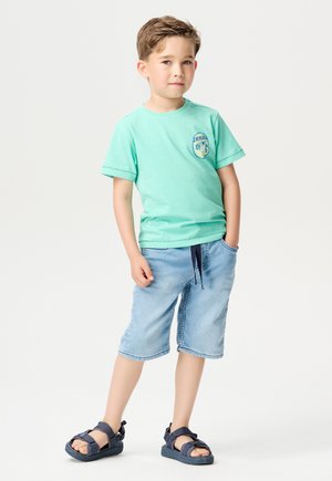 Hellgrünes Baumwoll-T-Shirt mit Logo, kombiniert mit hellblauen Denim-Shorts und dunkelblauen Sandalen, die über einen verstellbaren Kordelzug und Taschen verfügen.