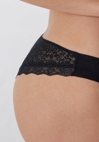 Schwarze Unterwäsche mit einem Spitzenbund und einem glatten Stoffkörper. Die Spitze hat ein florales Muster und einen gewellten Rand.
