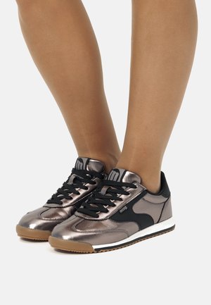Las zapatillas metálicas en bronce presentan un acento de tela negra, cordones negros, suela de goma y un diseño elegante con perfil bajo.