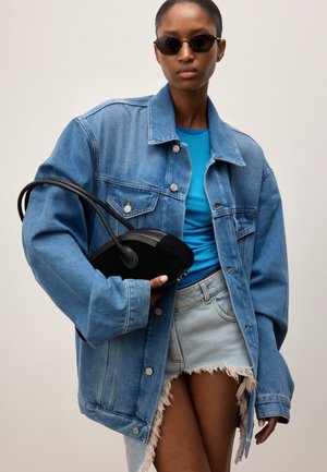 Femme portant une veste en jean bleue oversize, un haut bleu vif, une jupe en jean effilochée, des lunettes de soleil noires, tenant un sac à main noir sur un fond uni.