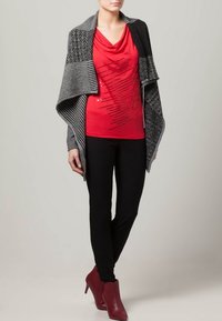 Svart och gråmönstrad kofta över en röd tröja med krage och grafisk design, matchad med svarta leggings och bordeauxfärgade ankelstövlar.