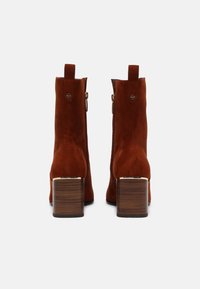 Bottines en daim marron avec un talon carré, dotées d'une fermeture éclair sur le côté et d'une petite languette à l'arrière pour faciliter l'enfilage.