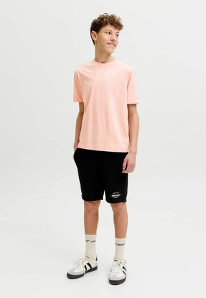 Adolescent garçon debout regardant à droite, portant un T-shirt rose clair, un short noir avec texte blanc, des chaussettes crème et des baskets blanches avec des rayures noires.
