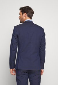 Marinblå kostymjacka med strukturerade axlar och slätt tyg. Har en enkel slits, långa ärmar och fyra knappar på varje manschett.