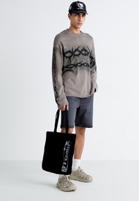 Pull gris avec un motif graphique noir, shorts gris, sac fourre-tout noir avec le logo "ATOMIC", et baskets beiges. Le modèle pose devant un fond uni.