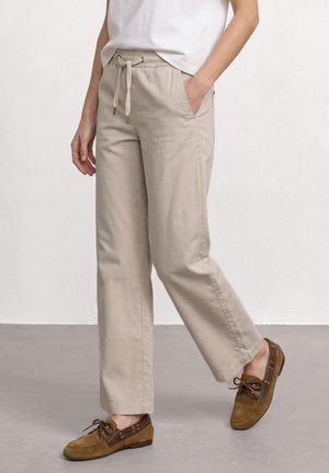 Pantalon beige à cordon porté avec des chaussures bateau en cuir et en daim marron sur une personne debout contre un mur clair.
