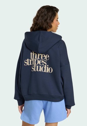 Vrouw met krullend haar, gekleed in een marineblauwe hoodie met de tekst "three stripes studio" en lichtblauwe korte broek, van achteren gezien tegen een lichte achtergrond.