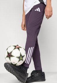 Pantalones deportivos morados con la marca de tres rayas blancas, diseño ajustado, combinados con zapatos de fútbol negros y un balón de fútbol con patrón.