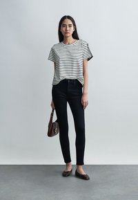 Femeie stând sprijinită de un perete simplu, purtând un tricou cu dungi albe și negre, blugi skinny negri, balerini maro și ținând o geantă maro.