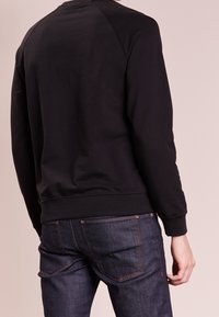 Černá mikina s kulatým výstřihem a manžetami a lemem s žebrováním. Tkanina vypadá hladce. Nosená s tmavě modrými džíny s oranžovými švy.