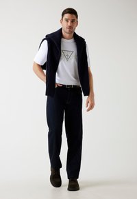 Hvid t-shirt med grafisk tryk, marineblå strikket vest, mørke denim bukser og brune fodtøj; afslappet outfit med lagdelt design og teksturer.