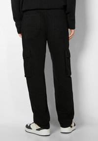 Bakifrån av en person som bär svarta cargo-byxor med lockiga fickor och svart-vita sneakers, som står mot en enkel bakgrund.