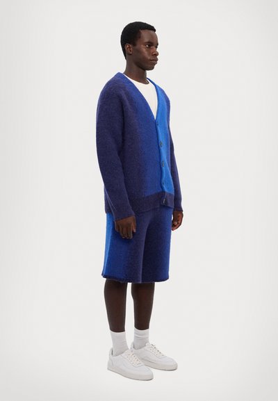 Blaue und lila strukturierte Strickjacke mit Knöpfen, kombiniert mit passenden Shorts und ergänzt durch weiße Sneaker und Socken.