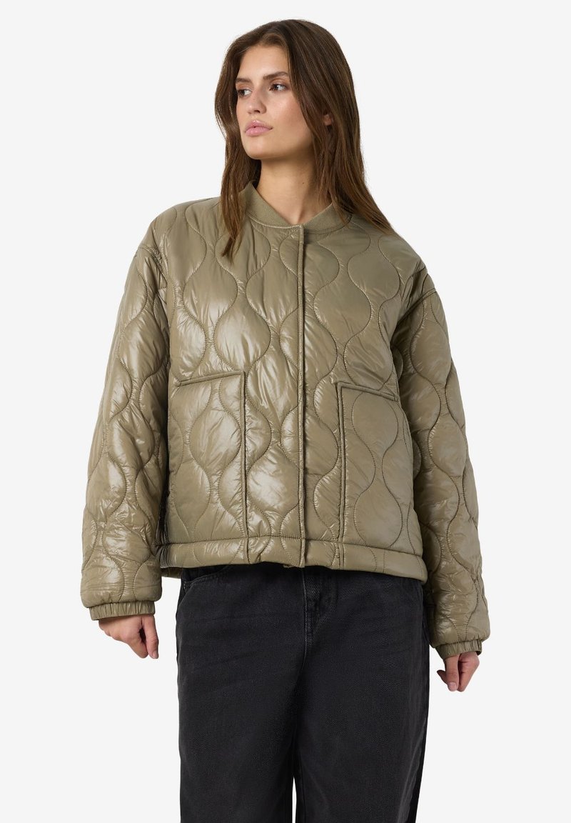 Γυναίκα που φοράει μπουφάν σε χακί πράσινο με κυματοειδή σχέδια και μεγάλες μπροστινές τσέπες, συνδυασμένο με μαύρο παντελόνι, κοιτώντας προς τα πλάγια.