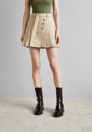 Vrouw die een beige geplooide rok met knopen en een riem draagt, een groene mouwloze top en donkerbruine enkellaarsjes op een licht tapijt.