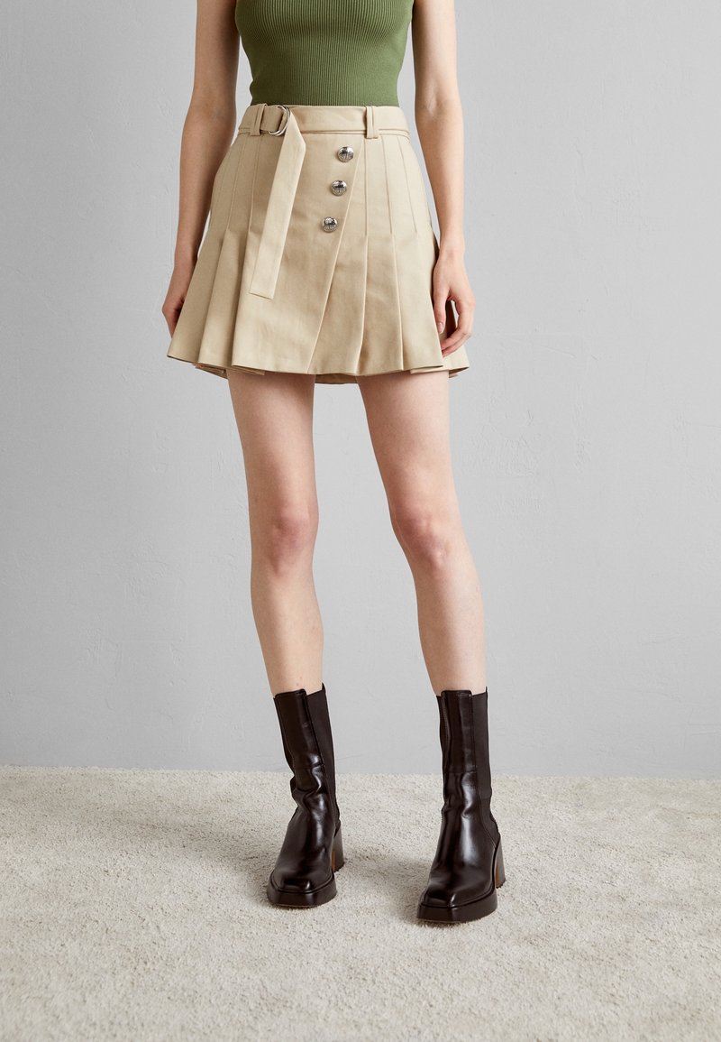 Femme portant une jupe plissée beige avec des boutons et une ceinture, un haut vert sans manches et des bottines marron foncé sur un tapis clair.
