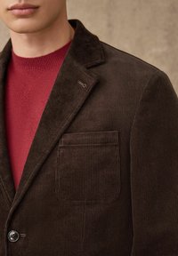 Hnědý manšestrový blejzr s klopovým límcem, zapínáním na jeden knoflík a přední kapsou. Nositý přes červený pletený svetr.