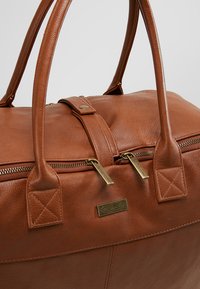 Borsa da viaggio in pelle marrone con manici doppi, zip dorate, logo "Kidzoom" in rilievo e dettagli trapuntati sulle attacchi dei manici.