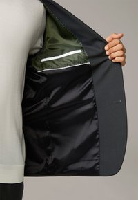 Giacca da costume su misura con tessuto esterno grigio, fodera di satin scuro e interno verde a contrasto con tasca con zip.