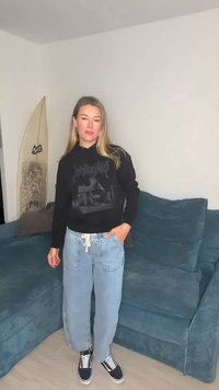 Crna majica s kapuljačom s grafičkim printom, uparena s svijetloplavim traper hlačama i tamnoplavim tenisicama. U pozadini plavi teksturirani kauč s daskom za surfanje.