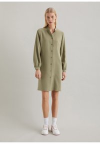 Abito verde oliva con bottoni, colletto alto, maniche lunghe a palloncino e tessuto testurizzato, che arriva a metà coscia, abbinato a sneaker bianche.