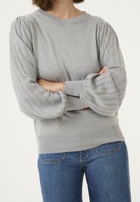 Pull gris clair avec manches ballon côtelées et col rond. Il présente un ourlet inférieur ajusté et est associé à un jean bleu.