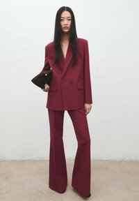 Vínově červený oblek na míru s dvouřadým sakem a rozšířenými kalhotami, vyrobený z hladké látky, držený s tmavým psaníčkem. Jednoduché pozadí.