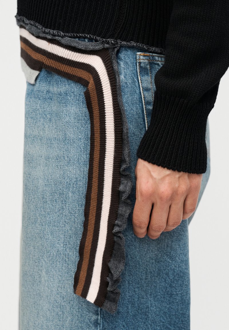 Main reposant sur le côté, portant un pull côtelé noir et un jean bleu avec un détail en tricot à rayures multicolores et un volant le long du bord de la poche.