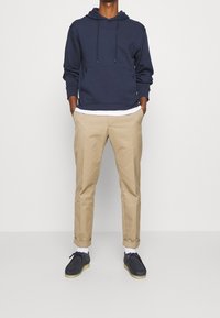 Man i marinblå hoodie, vit t-shirt under, beiga upprullade byxor, vita strumpor och svarta fritidsskor som står med händerna i fickorna.