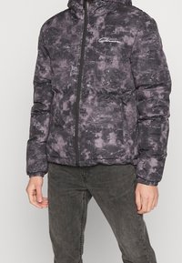 Man i en svart och grå mönstrad pufferjacka med dragkedja fram och mörkgrå jeans, stående mot en enkel bakgrund.