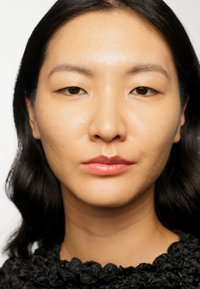 Nära bild av ett ansikte med slät, något glänsande hy som bär en topp av rynkat svart tyg, med fylliga läppar och rakt svart hår.