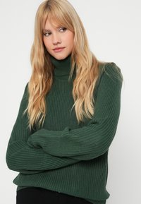 Suéter de gola alta verde em malha, com textura canelada, corte solto e mangas longas. Punhos e barra acentuados destacam um design oversized.