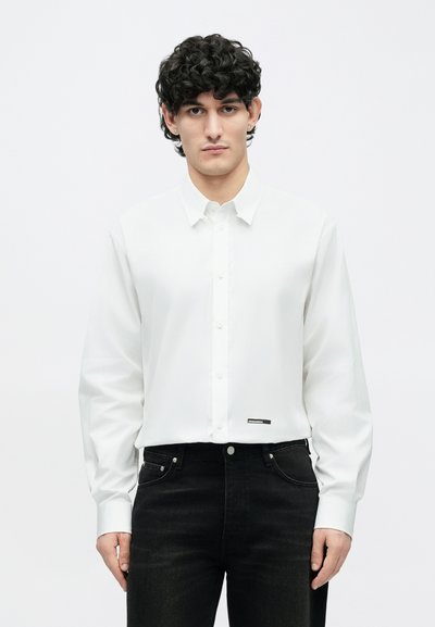 Mand med krøllet hår iført en hvid skjorte med knapper og sorte jeans, stående foran en ensfarvet hvid baggrund.